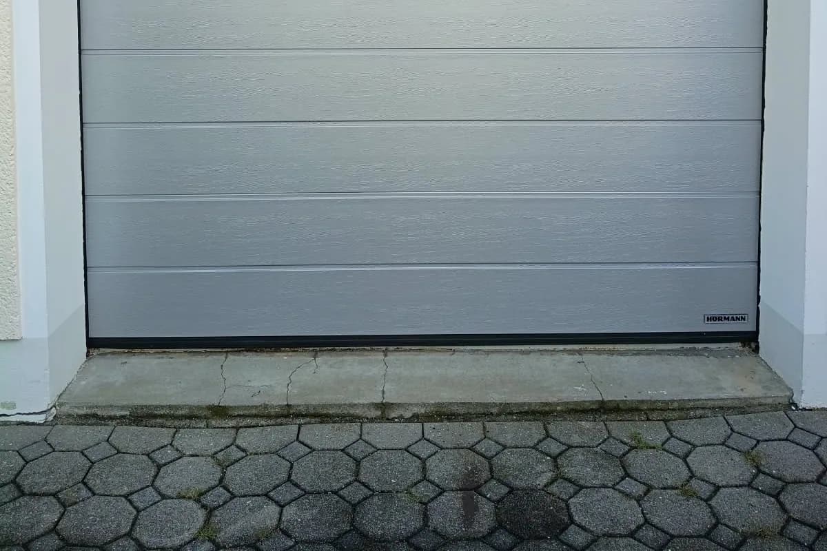 Rissige Betonschwelle vor einem grauen Garagentor mit angrenzendem Betonpflaster.